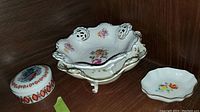 Three porcelain items on wooden shelf: footed oval decorative bowl, small scalloped bowl, round lidded container with figure and border