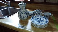Wide view of the set on wooden counter by sink showing teapot, cup, plates, and bowls with blue floral designs.