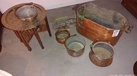 Overview photo showing folding carved wood base table with brass tray on top, large copper boiler, and several copper pots grouped together on basement floor.