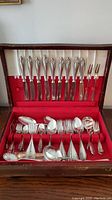 Full view of flatware set arranged inside open wooden box with red velvet and white satin interior lining showing knives, forks, spoons, and ladles.