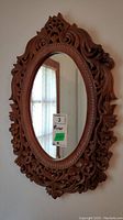 Full view of the oval mirror with ornate carved wood frame hung on wall, showing intricate wood carvings and reflective glass.