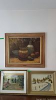 Three framed paintings displayed on wall: top a still life, bottom two landscape scenes