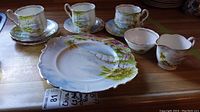 Tea set items displayed with cups, saucers, plate, creamer and sugar bowl on wooden surface, showing detailed hand-painted landscape design.