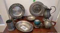 Photo showing full assortment of pewter items including plate, bowl, mug, candle holder, pitcher, wall art plaque, and teapot displayed on wooden cabinet against white wall.