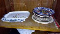 Photo of a collection of serving platters and stacked plates on a wooden shelf, showing a mix of decorated ceramic items including a rectangular floral Rosenthal platter and a white platter with blue floral border.