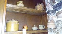 Shelf with all pottery items displayed including two pitchers, salt jar, three bowls with lids, and woven tray on top shelf