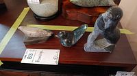 Photo of three soap stone carvings on wooden base - includes a bird, a human figure, and a broken carving. Shows lot condition and arrangement.