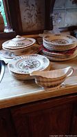 Wade Meadow dinnerware set displayed on wooden surface, includes stacked plates, bowls, two lidded serving bowls, and gravy boat in front.