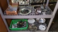 Wide shelf view including escargot utensil box, silver-plated trays, pewter plates, and a green basket of flatware below.