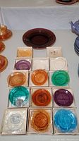 Overview of collection of 18 glass cup plates arranged on a white cloth background including multiple colors and some still boxed.