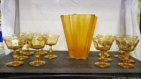 Front view showing vintage amber glass vase with fluted design, surrounded by eight amber glass stemmed glasses on black surface.