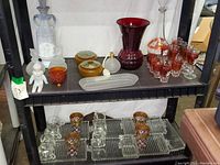 Shelf display of assorted vintage glassware including clear and red cordials, a red vase, clown figurine, perfume bottles, appetizer trays and decanter.