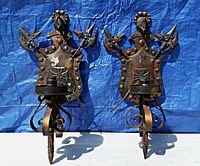 Front view of two cast iron wall light fixtures displaying coat of arms motifs with shield, spears, rivets, and finials on a blue tarp background for detail.
