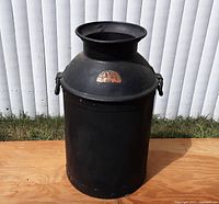 Full front view of antique metal milk can painted black with visible wear and a plaque on the front.