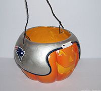 Side angle showing orange pumpkin candy pail with a metallic silver football helmet featuring New England Patriots logo on the helmet's side and a carved jack-o-lantern smiling face on the pumpkin.