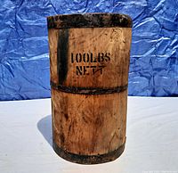 Photo of the wooden nail barrel showing the front marking '100 LBS NET T' with metal hoops around the top, middle, and bottom, clear weathered wood grain and some stain marks.