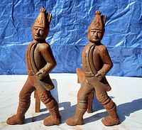 Front view of two rusted cast iron andirons shaped as Hessian soldiers with detailed hats and sashes, standing upright.