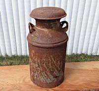 Front view of rusty metal milk can with embossed 'LYNDONVILLE CREAMERY' lettering, lid on top, two handles on neck, placed outdoors against white fence on wooden plank.