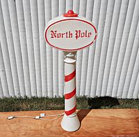 Front view of the white and red plastic North Pole sign lawn decoration standing upright on wood, with the 'North Pole' text visible on the oval sign area.
