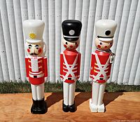 Front view of all three plastic blow mold Christmas yard decorations: one nutcracker and two soldiers standing side by side on wooden surface against white siding background.