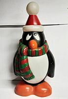 Front view of vintage penguin blow mold showing silly expression, red Santa hat with white trim and pom-pom, red and green striped scarf, orange beak and feet, and black and white body.