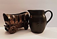 View of covered wagon-shaped planter with shiny metallic glaze and black cowboy pitcher side by side
