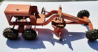 Side view of vintage orange metal toy road grader showing overall length and detailing, including cab area, wheels, and grading blade.