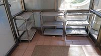 Three metal and plastic boot trays/shelves with rust on frames, set in an outdoor/function room area with tiled floor and large windows.