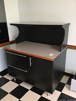 Front side view of black laminate cabinet with raised top shelf showing two drawers and cabinet door with metal handles and metal legs.
