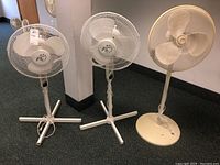 Photo showing three standing electric fans: two white FC brand and one beige, arranged side by side on carpeted floor.