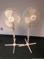 Two white Hampton Bay standing fans are shown side by side against a wall, both have tripod bases and corded power supply. Fans are similar in height and design.