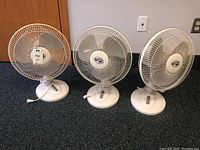 Three white Hampton Bay table fans lined up on the floor, visible from front showing grills, bases, and power cords.