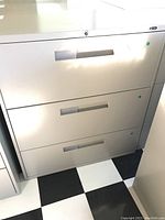Front view of white metal filing cabinet with three drawers and horizontal integrated handles, showing drawer fronts with minor scratches.