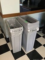 Image showing two gray plastic garbage cans side by side on a black and white checkered floor. The left garbage can has a clear plastic liner bag.