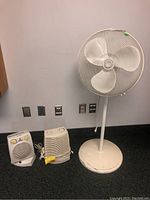 Three white appliances: two space heaters and one pedestal floor fan against a wall with power outlets.