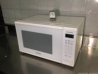 White Panasonic microwave model NN-SG62W with refrigerator thermometer placed on top, front view showing control panel and door.