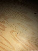 Close-up photo of light-colored plywood showing natural wood grain and a metal nail embedded on surface.