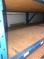 Plywood sheets stacked on blue metal shelving against a painted brick wall