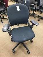Front view of black Steelcase office chair showing cloth seat with stains and black vinyl arms on caster wheels.