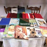 Top-down view of all books laid out on a white cloth on table and chairs visible in background. Shows variety of books including dog books, atlases, red and green hardcovers, and Antique Roadshow book.