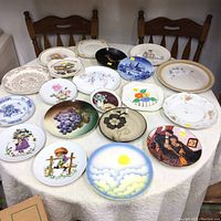 Photo of all 18 decorative collector plates arranged on a round table with a white tablecloth, showing a variety of floral, scenic, and artistic plate designs.