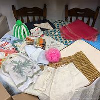 Wide view of the collection showing various placemats, a knitted tea cozy, dish towels, bath pouf, and other textiles laid out on chairs and surface.