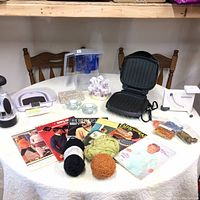 Full lot on round table showing kitchen appliances, knitting books and yarn, candle holders, and other small household items.