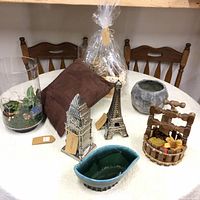 Overview of all items on white round table: metal statues of Big Ben and Eiffel Tower, glazed green bowl, glass terrarium, small grey bowl, wooden fountain sculpture, brown cloth, wrapped decorative basket