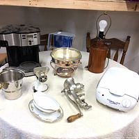 Full view of kitchen items on white tablecloth, showing coffee maker, water pitcher, meat grinders, chafing dish, gravy boat, and lamp.