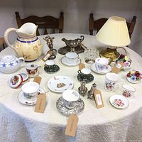 Full view of the collection on a round table covered with a white tablecloth showing ceramic pitcher, cups, saucers, lamp, candle holders, and porcelain figurines.