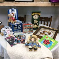 Full view of all items on a table including chair with teddy bear, plush rabbit, comic books, green clock with teddy bear, framed dog picture, handmade doll, decorative items, and game container.