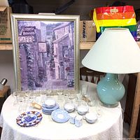 Full assortment on round table: framed cafe print, ceramic lamp, coffee glasses, tea sets, and decorative plate