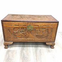 Front view of the wooden chest showing detailed hand carved panel depicting a scene with figures and structures, round metal lock plate present but missing clasp.