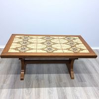 Front view of rectangular coffee table with beige and brown geometric tile top bordered by wood frame on light wood floor against plain wall.